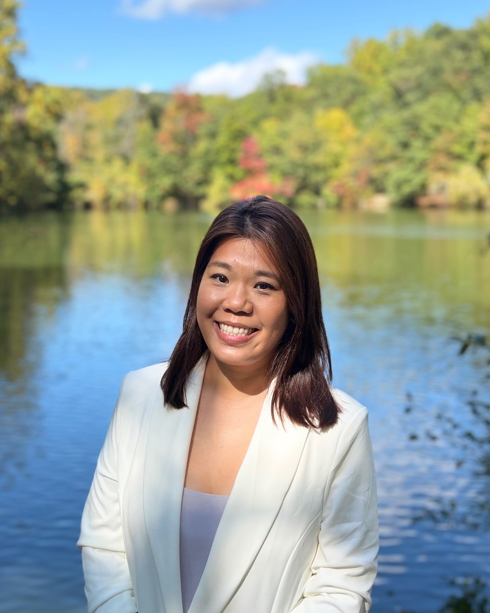 Jennifer Russ smiling in a white blazer beside a lake with autumn trees